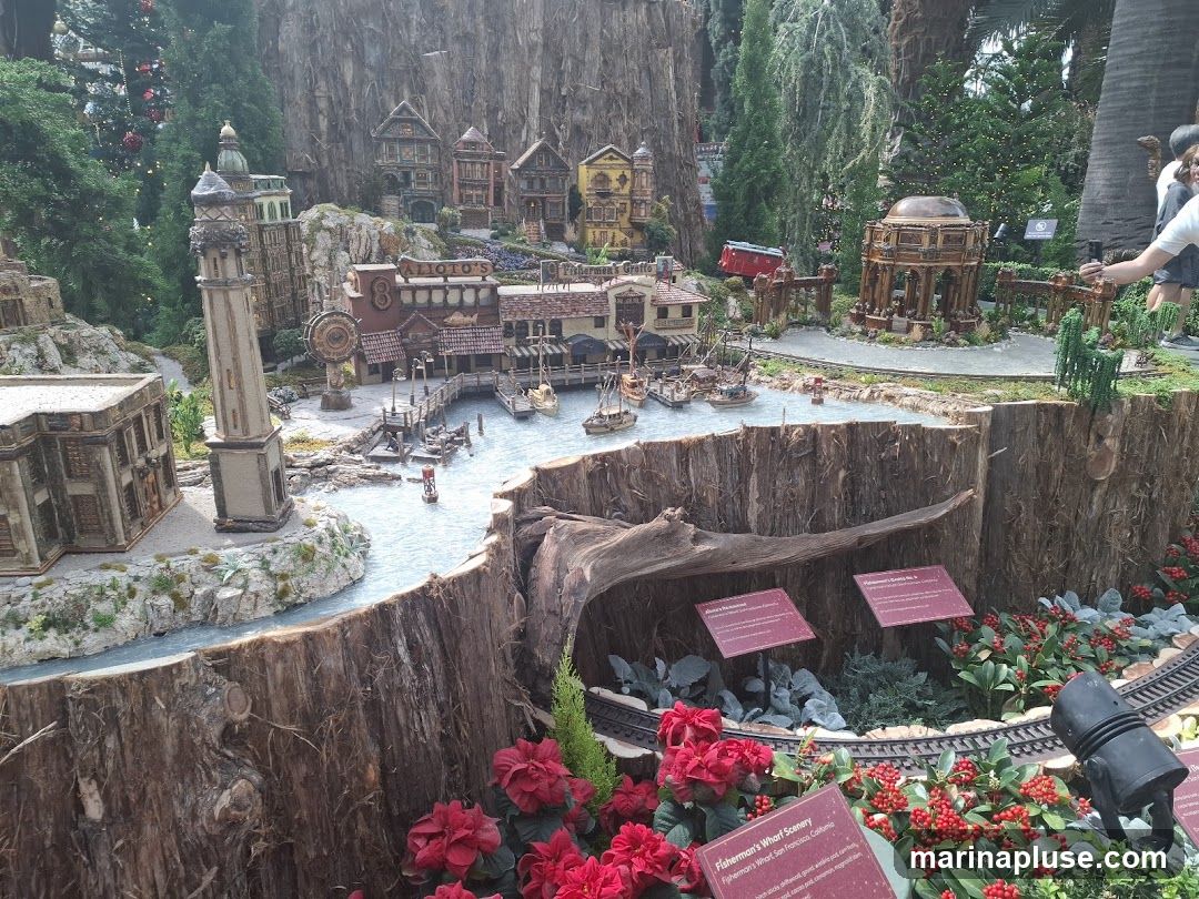A detailed miniature diorama depicting a bustling Fisherman's Wharf scene with boats in a harbor, various buildings, a lighthouse, and a train track. The diorama is set against a backdrop of trees and rocks, with red flowers and greenery in the foreground. Informational signs are placed around the display.