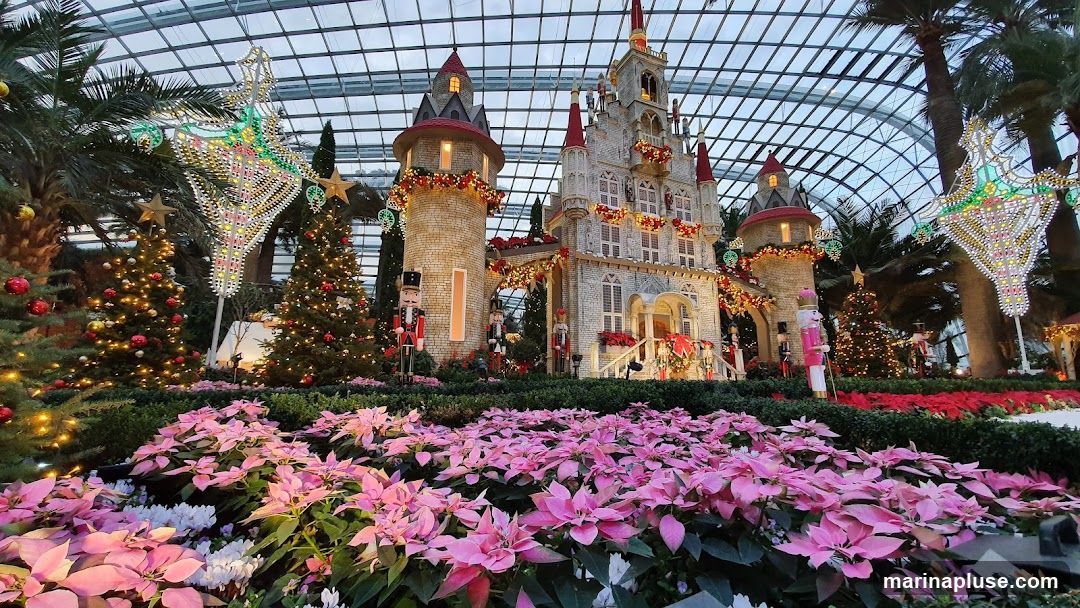 A festive Christmas display featuring a large, ornate castle with towers, decorated with garlands and lights. In front of the castle, there are Christmas trees adorned with ornaments and lights, as well as nutcracker figures. A foreground of pink and white poinsettias and other greenery completes the scene, all under a glass-domed ceiling.