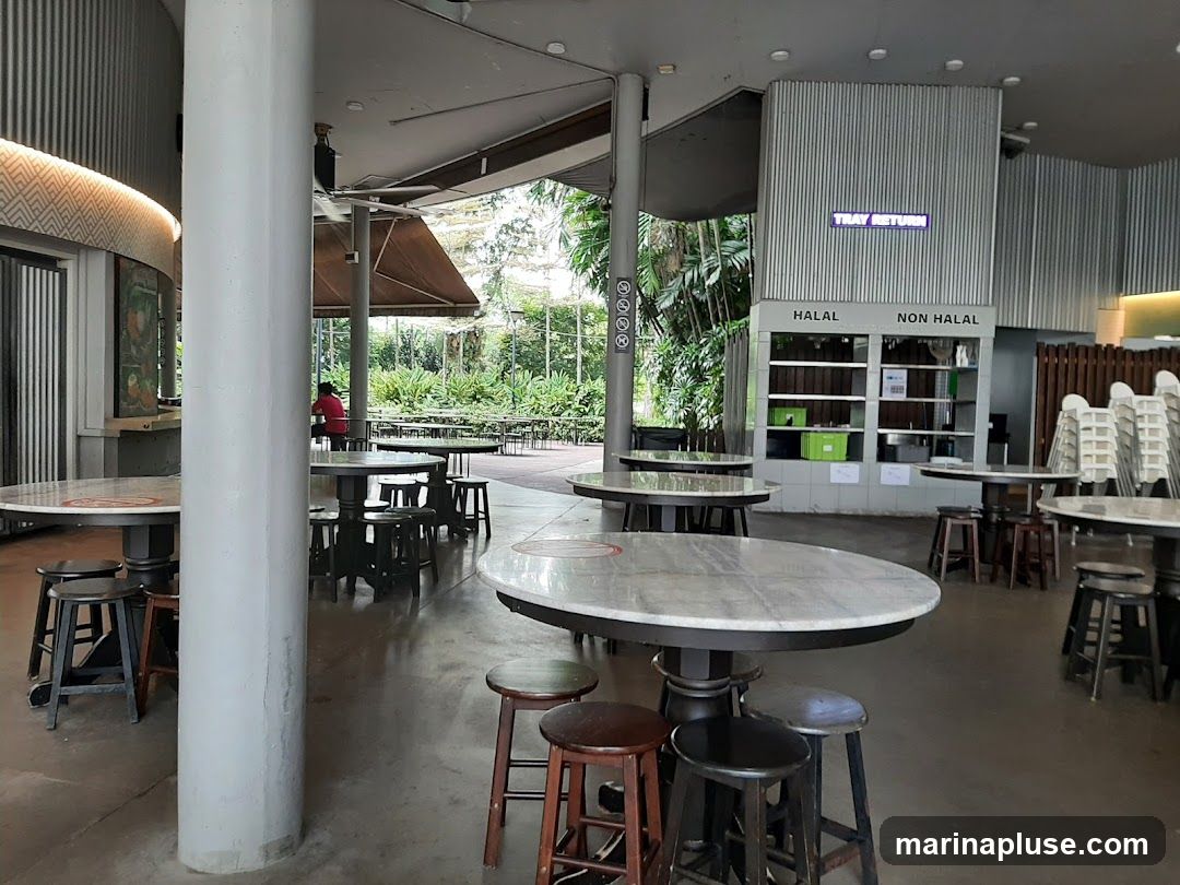 An empty food court with round marble-topped tables and stools. In the background, there is a tray return station with "HALAL" and "NON HALAL" sections. Stacks of chairs are visible to the right. Lush greenery can be seen through an open-air section of the food court.