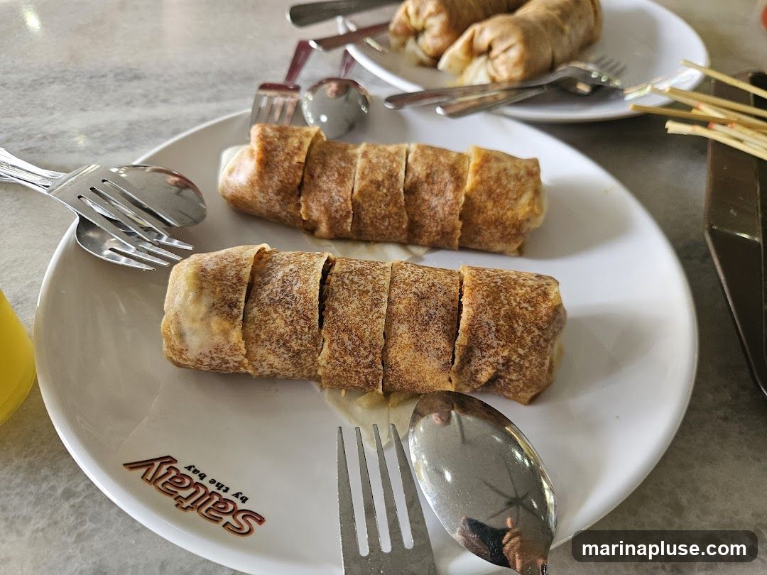 Two spring rolls, sliced into segments, are presented on a white plate. The plate has a logo that reads "Satay by the Bay". Cutlery, including forks and spoons, is visible around the plate. Another plate with spring rolls is in the background.