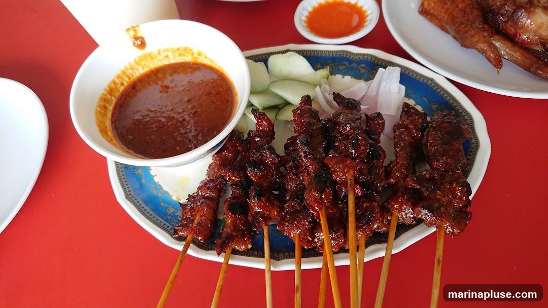 A plate of grilled satay skewers served with a bowl of peanut sauce, sliced cucumbers, and onions. A small dish of chili sauce is also visible, along with a portion of roasted chicken on another plate in the background. The food is arranged on a red table.