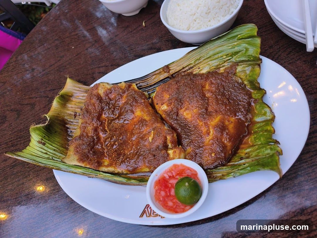 Two pieces of grilled fish, coated in a rich, reddish-brown sambal sauce, are served on a bed of grilled banana leaves. A small white bowl contains a side of chopped tomatoes, onions, and a lime wedge. A bowl of white rice is visible in the background, along with stacked white plates and chopsticks. The meal is presented on a wooden table.