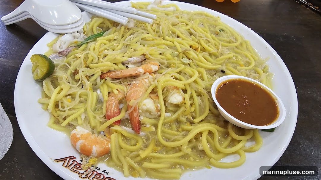 A plate of yellow noodles with shrimp, squid, and a side of dipping sauce. A lime wedge is also visible on the side of the plate. The plate has a logo that reads "Sattay".