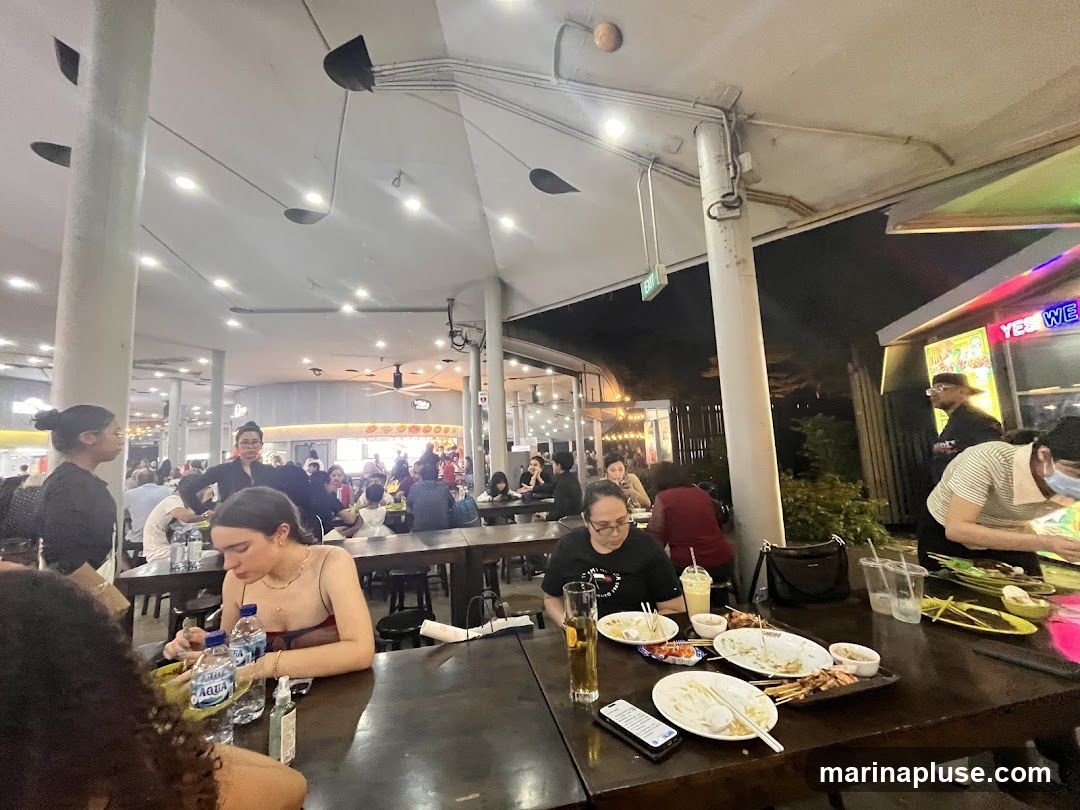 A busy outdoor food court with many people seated at tables, eating and socializing. The lighting suggests it is evening. In the background, there are various food stalls and a neon sign that says "YES WE". The tables are laden with plates of food, drinks, and used utensils.
