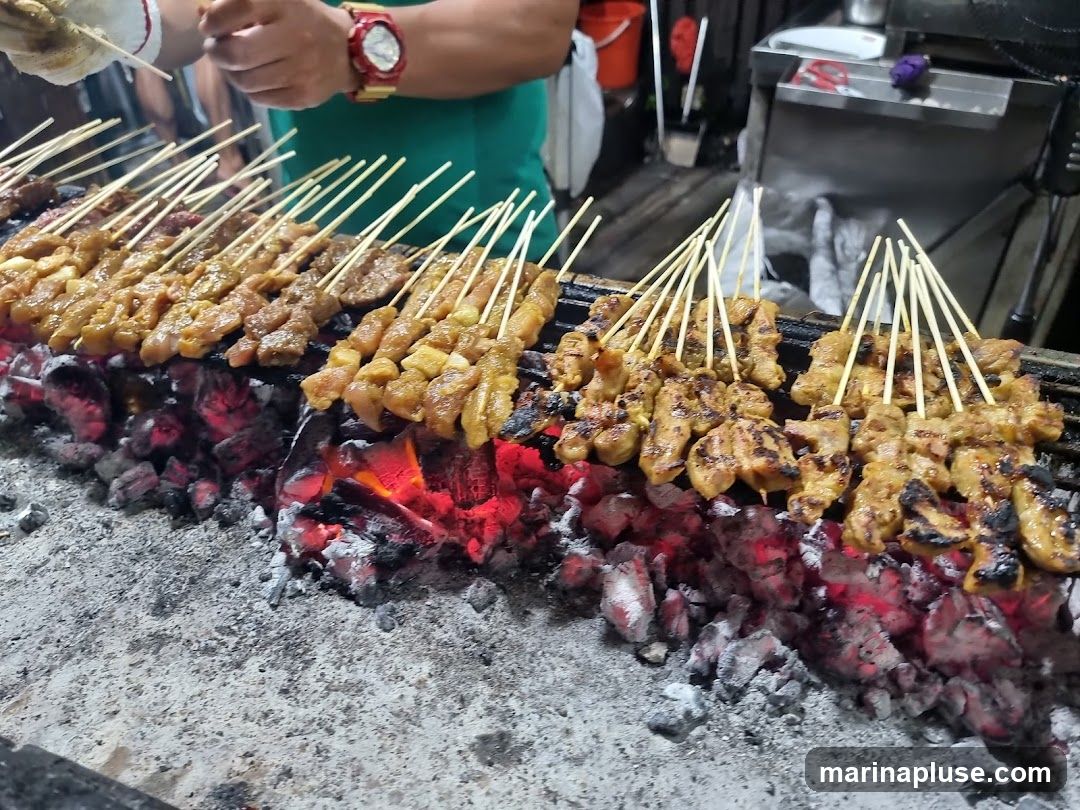 A close-up shot of numerous skewers of marinated meat, likely sate, being grilled over glowing red coals. The meat appears to be chicken and possibly pork, with some pieces showing char marks from the grill. A person wearing a green shirt and a watch is visible in the background, tending to the skewers. The scene suggests a street food vendor or a casual eatery.