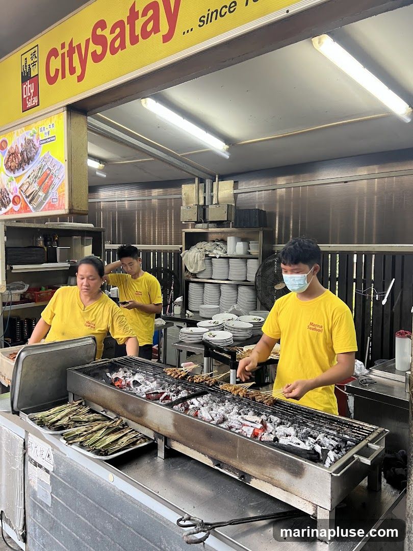 Three people in yellow shirts are working at a food stall. Two are preparing satay over a charcoal grill, and one is standing behind them. A sign above the stall reads "CitySatay ... since 1978". There is a menu board with pictures of satay dishes.