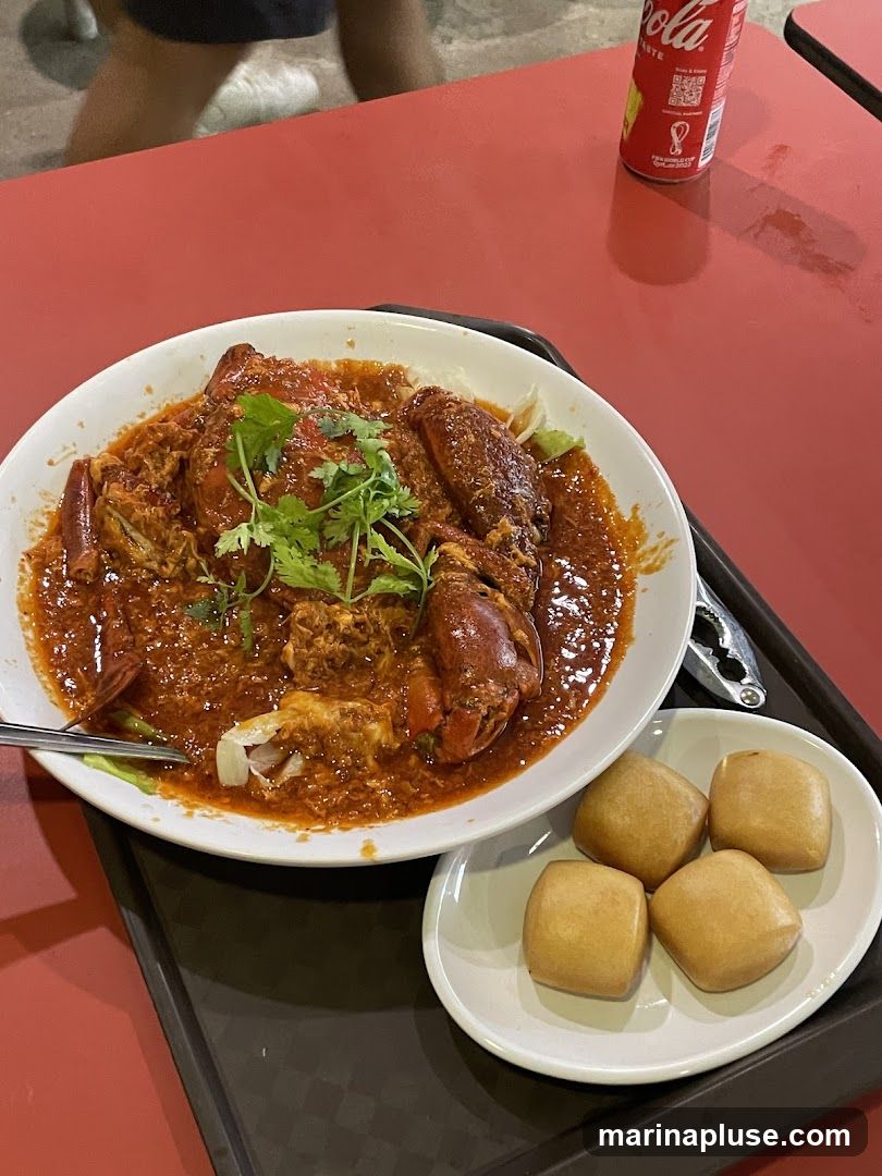A plate of chili crab served with a side of fried mantou buns sits on a red table next to a can of Coca-Cola. The crab is coated in a thick, savory sauce and garnished with cilantro. The mantou buns are golden brown and appear to be deep-fried.