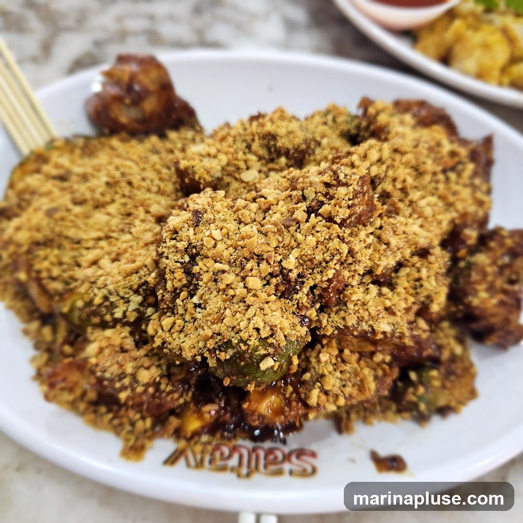 A close-up shot of a plate of rojak, a Malaysian fruit and vegetable salad, generously topped with crushed peanuts. The rojak appears to be a mix of fruits and vegetables coated in a dark, sweet and savory sauce. The plate has the word "satay" printed on it. In the background, another dish with a small bowl of sauce is visible.