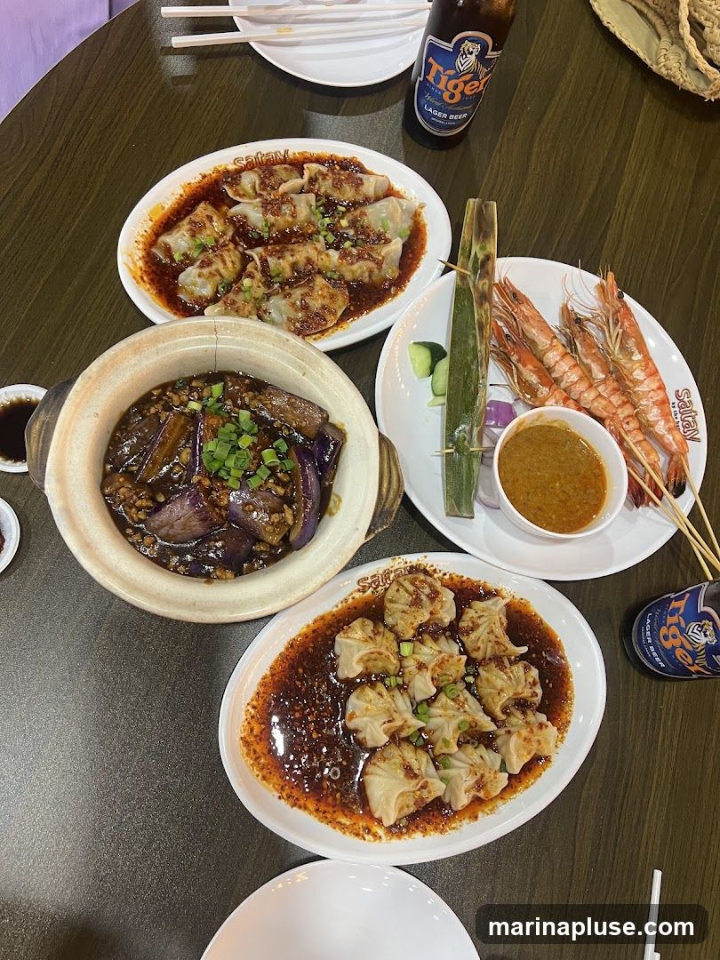 A top-down view of a table laden with various dishes and drinks. In the center, a clay pot holds a serving of braised eggplant with chopped green onions. To the left, a plate of dumplings in a spicy sauce is visible. To the right, a plate of grilled prawns on skewers is accompanied by a small bowl of dipping sauce and cucumber slices. Another plate of dumplings in a similar spicy sauce is positioned at the bottom of the frame. Two bottles of Tiger Lager Beer are also on the table, along with white plates and chopsticks. The overall impression is a shared meal with diverse culinary offerings.