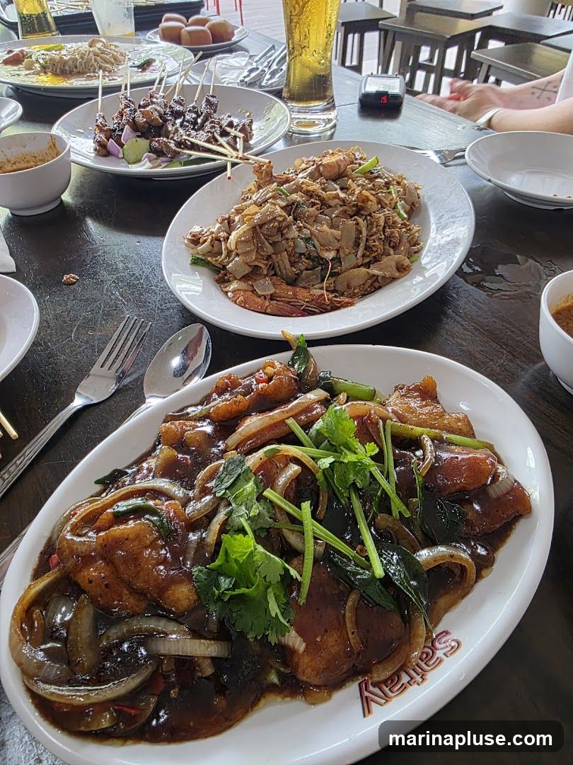 A table laden with several plates of food, including satay skewers, noodles with shrimp, and a dish with fish or meat in a dark sauce, garnished with onions and cilantro. A glass of beer and bowls of dipping sauce are also visible. The setting appears to be an outdoor or semi-outdoor dining area with wooden tables and chairs.