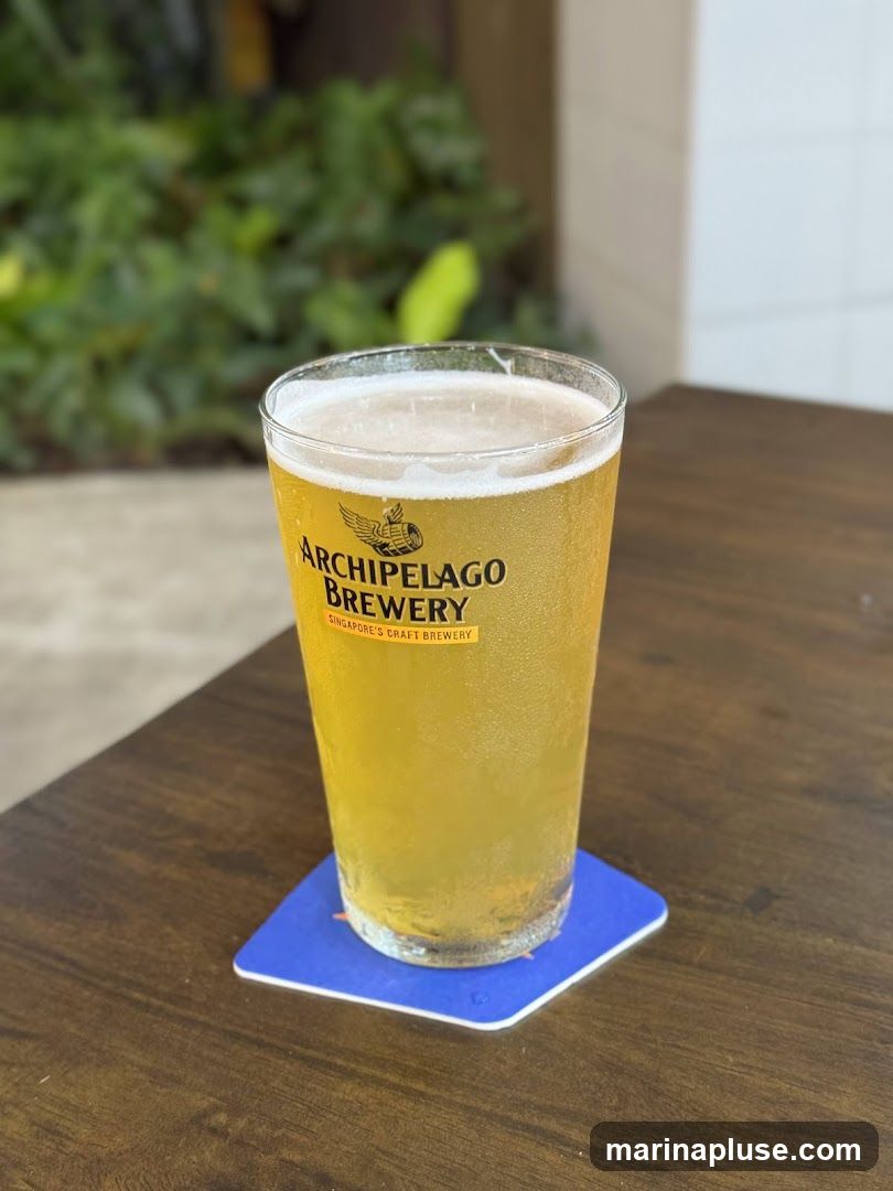 A close-up shot of a glass of beer with a foamy head, sitting on a wooden table. The glass has a logo that reads "ARCHIPELAGO BREWERY SINGAPORE'S CRAFT BREWERY". The background is blurred, showing greenery and a white tiled wall.