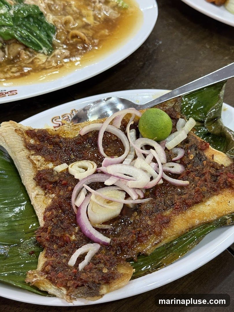 A close-up shot of a grilled fish dish served on a banana leaf. The fish is topped with a spicy chili paste and garnished with sliced red onions and a lime wedge. In the background, another dish with noodles and greens is visible.