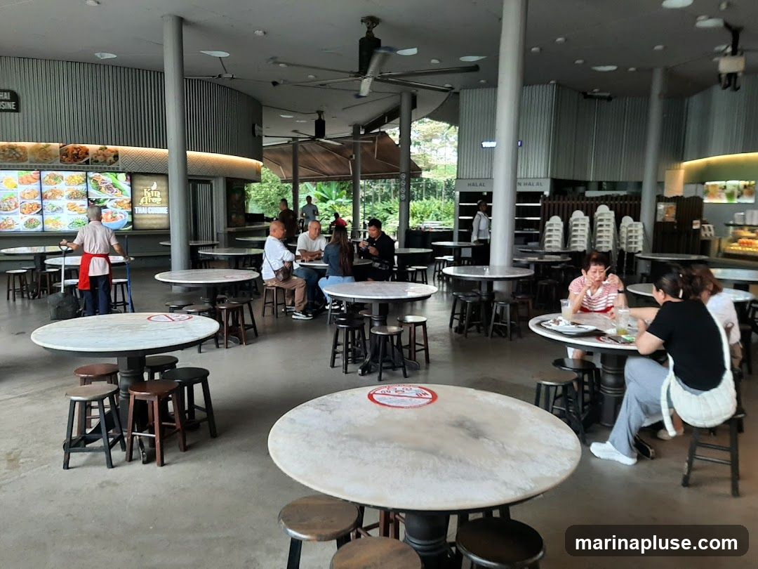 An open-air food court with multiple round tables and stools. People are seated at some tables, eating and conversing. A food stall with a menu board displaying various dishes is visible in the background. The overall ambiance is casual and bustling.