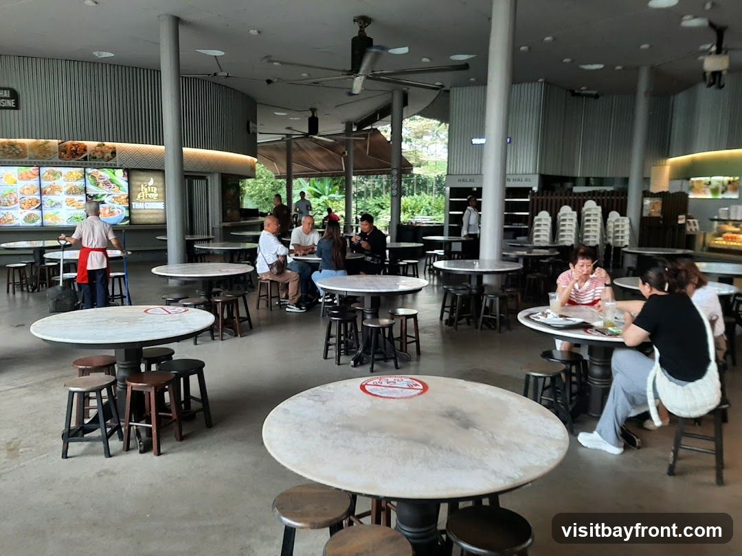 An open-air food court with multiple round tables and stools. People are seated at some tables, eating and conversing. A food stall with a menu board displaying various dishes is visible in the background. The overall ambiance is casual and bustling.