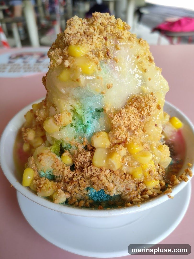 A close-up shot of a colorful shaved ice dessert. The dessert is topped with corn kernels, crushed peanuts, and a sweet syrup. The shaved ice has layers of blue and green, with a white ice cream scoop on top. The dessert is served in a white bowl on a pink table. In the background, there are blurred images of tables and chairs, suggesting an outdoor seating area.