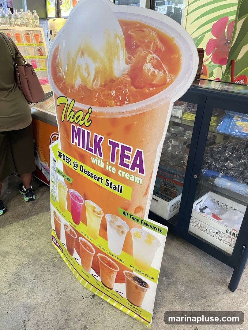A large cardboard standee displays a picture of Thai Milk Tea with ice cream and a list of various drinks. The standee is placed in front of a dessert stall.