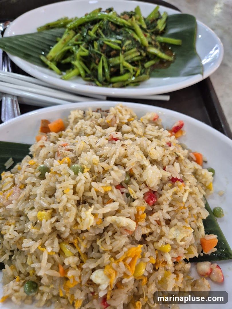 A close-up shot of two plates of food. The foreground plate is filled with fried rice containing peas, corn, carrots, and scrambled egg. The background plate holds a serving of stir-fried water spinach.