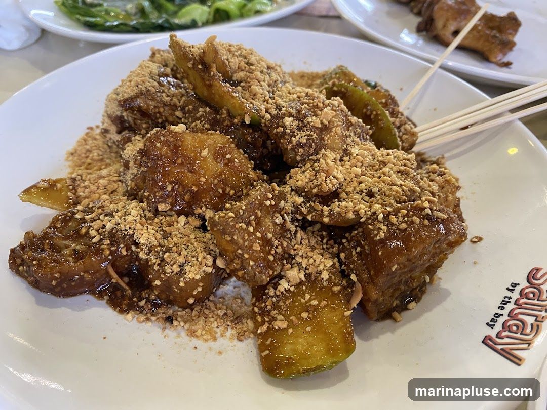 A close-up shot of a plate of rojak, a Malaysian fruit and vegetable salad, generously topped with crushed peanuts and a dark, sweet sauce. The rojak appears to be a mix of fruits and possibly some vegetables, cut into bite-sized pieces. The plate has a logo that reads "Salay by the bay". In the background, other dishes are visible, including a plate of green vegetables and a skewer of grilled food.
