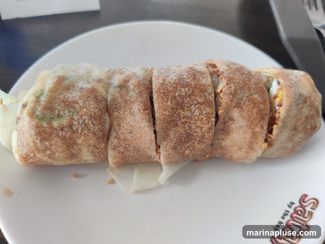 A close-up shot of a sliced spring roll on a white plate. The spring roll appears to be filled with various ingredients and is golden brown in color. The plate has a logo that reads "Saitai by the bay" in orange and white lettering.