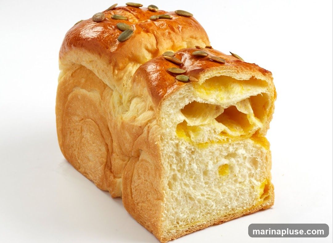 A loaf of baked bread with a golden-brown crust topped with pumpkin seeds, cut to reveal a soft, airy interior with yellow filling or swirls.