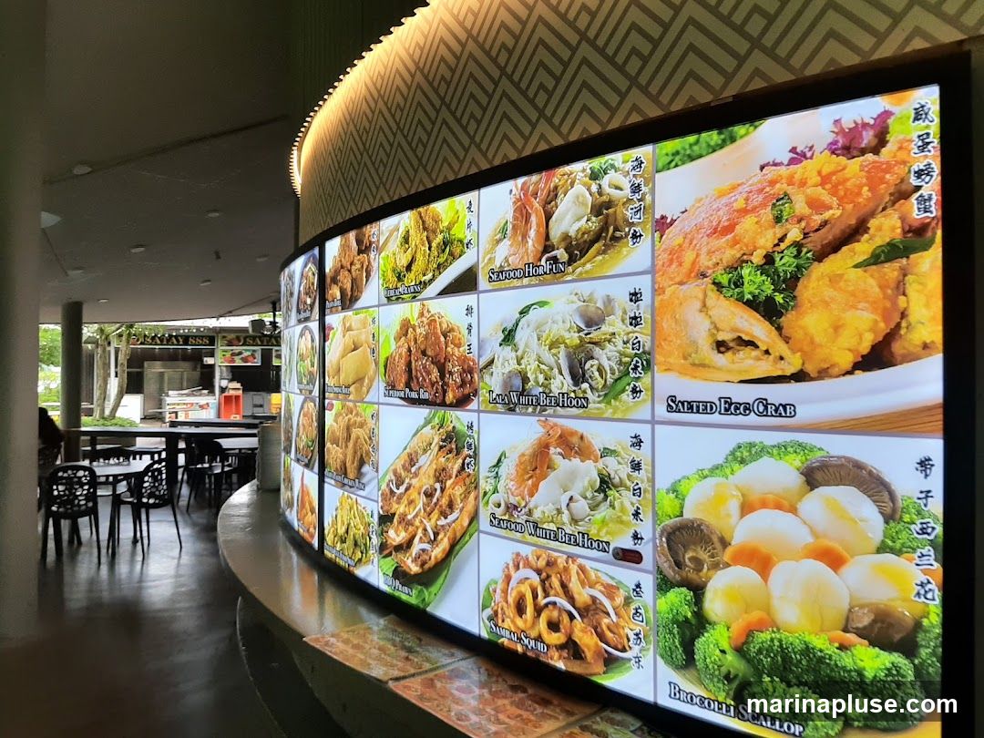 A large, curved digital menu board displaying various food items with images and names. The menu includes dishes like Salted Egg Crab, Seafood Hor Fun, Lala White Bee Hoon, Seafood White Bee Hoon, Sambal Squid, and Broccoli Scallop. In the background, a food stall labeled "SATAY 888" is visible, along with tables and chairs.
