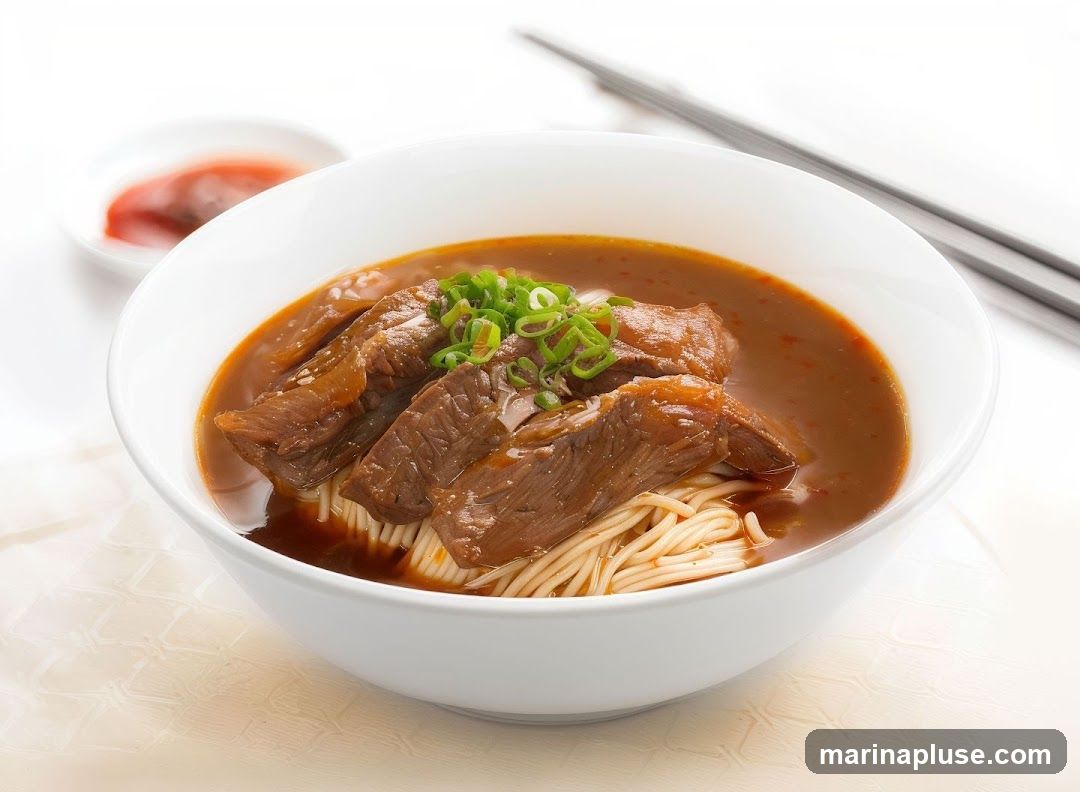 A close-up shot of a bowl of beef noodle soup. The soup is a rich brown color, topped with tender slices of braised beef and garnished with chopped green onions. Thin noodles are visible beneath the beef, and a small dish of red sauce is in the background. Chopsticks rest on the side.