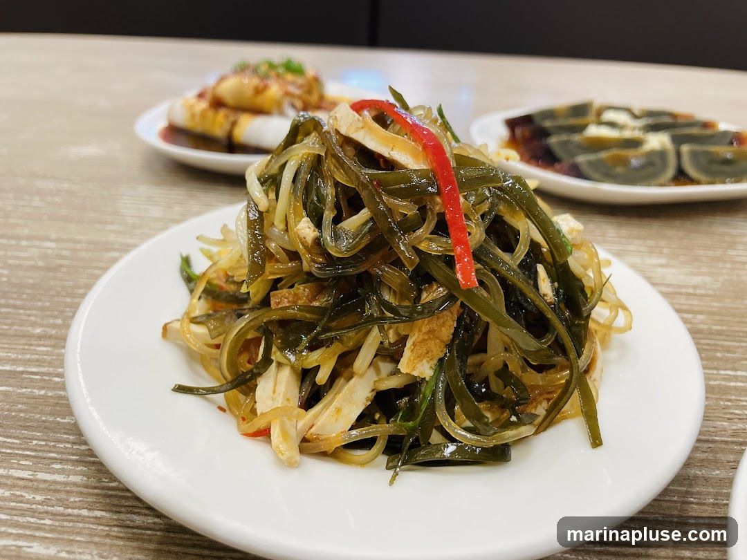 A close-up shot of a plate of seaweed salad, garnished with julienned red pepper and thin strips of tofu. In the background, two other plates of food are visible: one with what appears to be spring rolls topped with sauce and scallions, and another with sliced century eggs. The dishes are arranged on a wooden table.