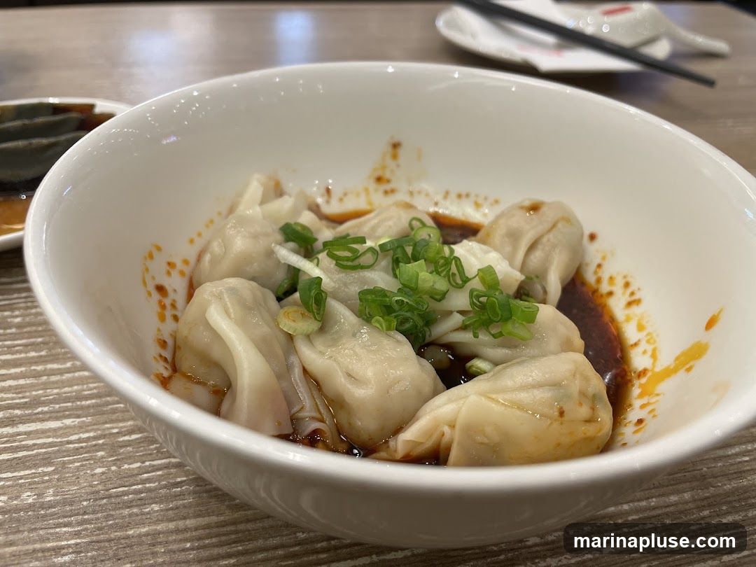 A close-up shot of a bowl of wontons in a spicy chili oil sauce, garnished with chopped green onions. In the background, a small dish of what appears to be preserved eggs and chopsticks on a plate are visible.