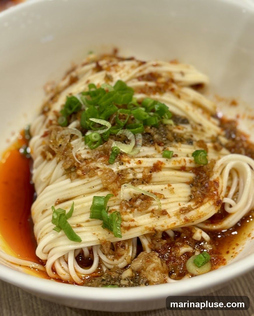 A close-up shot of a bowl of noodles, likely a spicy Sichuan dish, topped with chopped green onions and a flavorful sauce. The noodles are thin and white, arranged in a neat pile, and coated in a reddish-brown sauce with visible chili flakes and minced garlic. The dish is served in a white ceramic bowl.