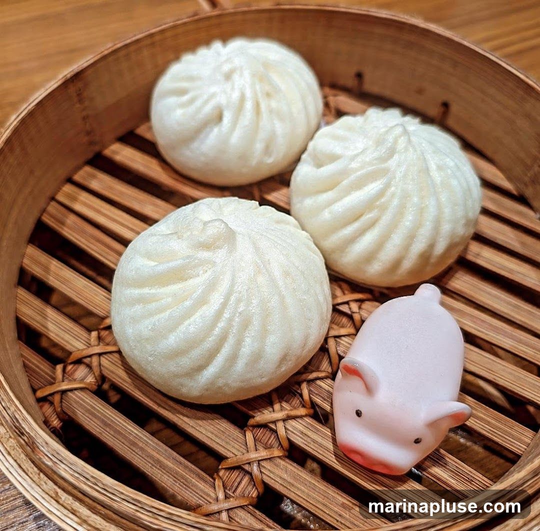 Three white steamed buns are arranged in a bamboo steamer basket. A small, pink, pig-shaped decoration sits next to the buns. The buns have a pleated top, characteristic of baozi. The steamer basket is placed on a wooden surface.
