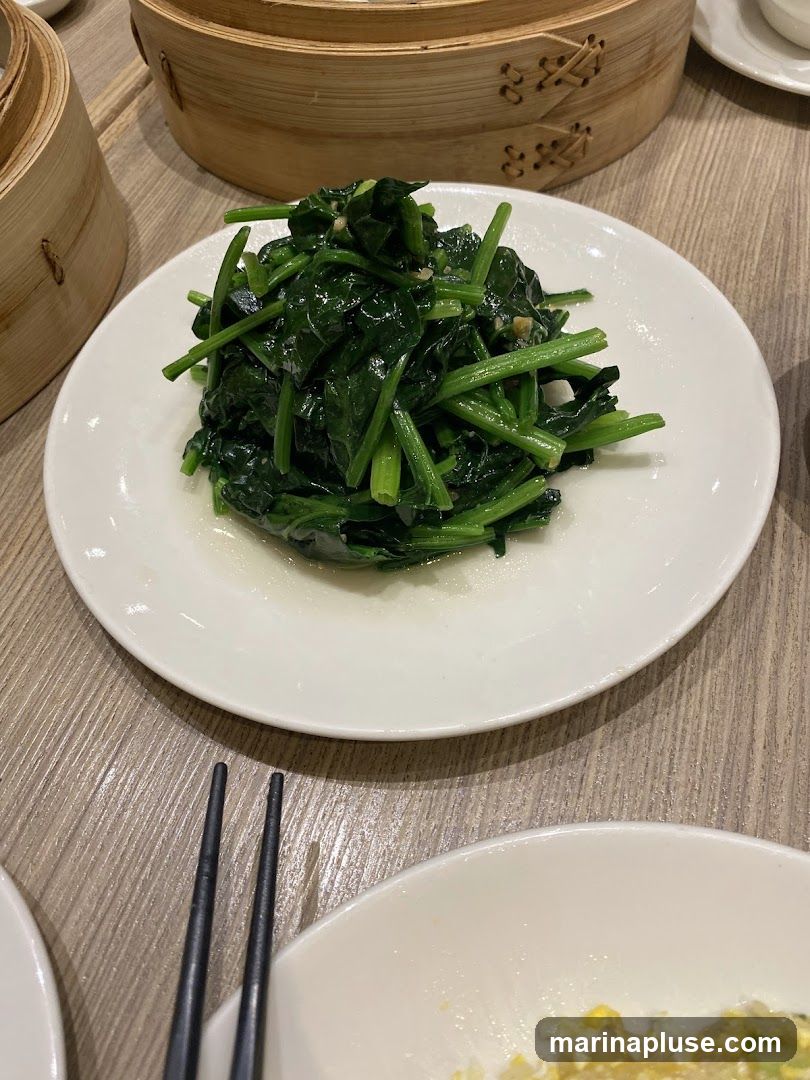 A plate of sautéed spinach with garlic is presented on a wooden table, with bamboo steamers in the background. Chopsticks are visible in the foreground.