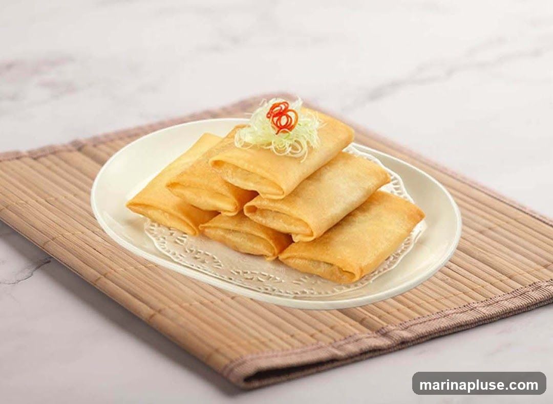 A plate of golden-brown spring rolls garnished with shredded scallions and red chili. The plate is set on a bamboo placemat with a marble-patterned background.