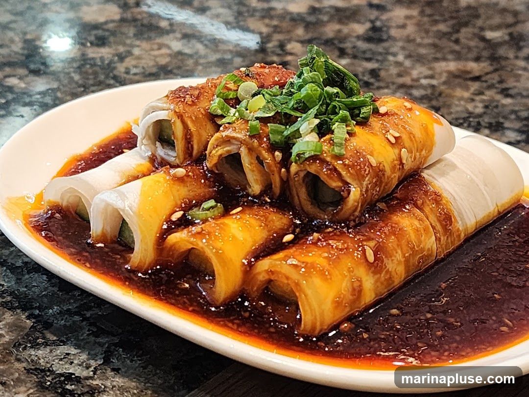 A white oval plate holds several rolled appetizers, possibly made with thin slices of tofu skin or a similar wrapper, filled with cucumber or another green vegetable. The rolls are coated in a glossy, dark reddish-brown sauce and sprinkled with sesame seeds and chopped green onions. The plate is set on a dark, speckled countertop.
