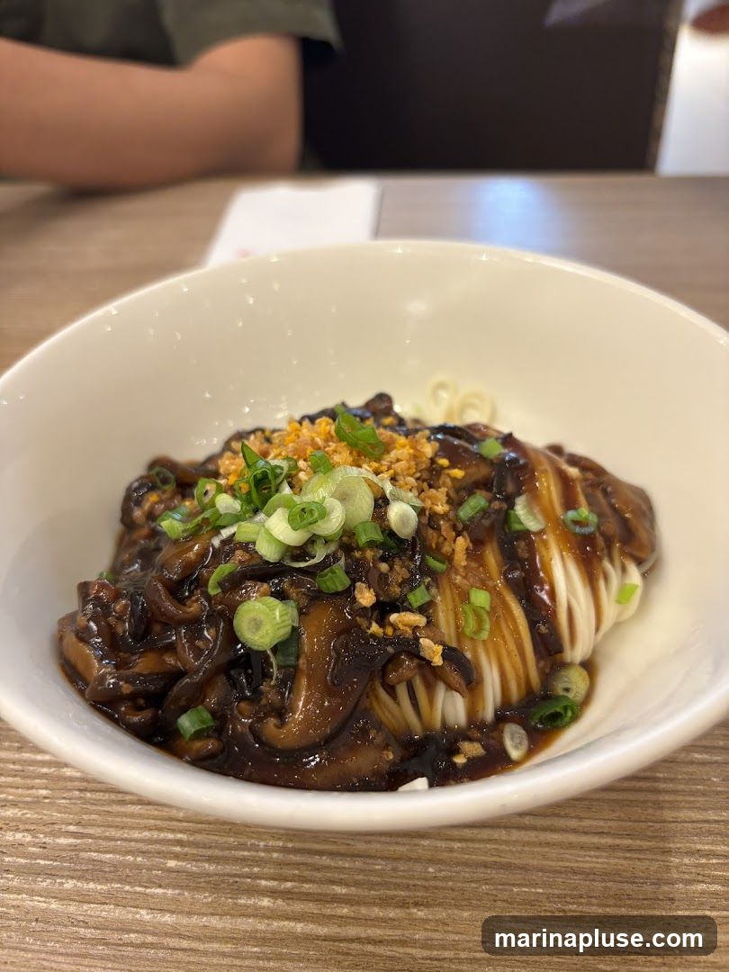 A close-up shot of a bowl of noodles topped with a dark sauce, sliced mushrooms, chopped scallions, and crispy fried garlic bits. The noodles appear to be thin and white, with the sauce generously coating the mushrooms. The dish is presented in a white bowl on a wooden table. A person's arm and shoulder are visible in the blurred background.