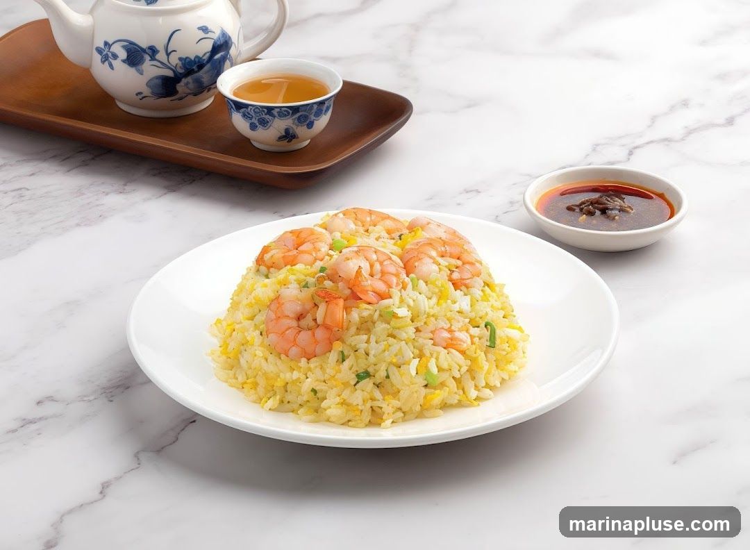 A plate of shrimp fried rice is served with a small bowl of chili sauce and a teapot with a cup of tea. The fried rice is topped with several large shrimp and garnished with chopped green onions. The dish is presented on a white plate with a marble-patterned background.