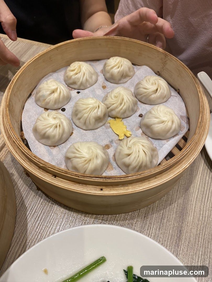 A bamboo steamer basket filled with ten soup dumplings, each with delicate pleats. A small yellow garnish is placed among the dumplings. In the foreground, a white plate holds some green vegetables. Two people's hands are visible in the background, suggesting a dining setting.