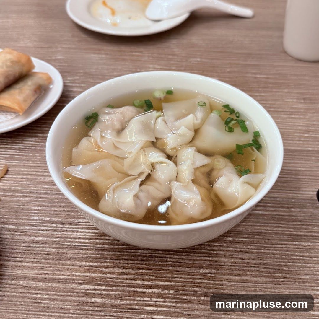 A close-up shot of a bowl of wonton soup, garnished with chopped green onions, sits on a wooden table. To the left, a small white plate holds two golden-brown spring rolls. In the background, out of focus, is another plate with a spoon and some food, and a white mug.