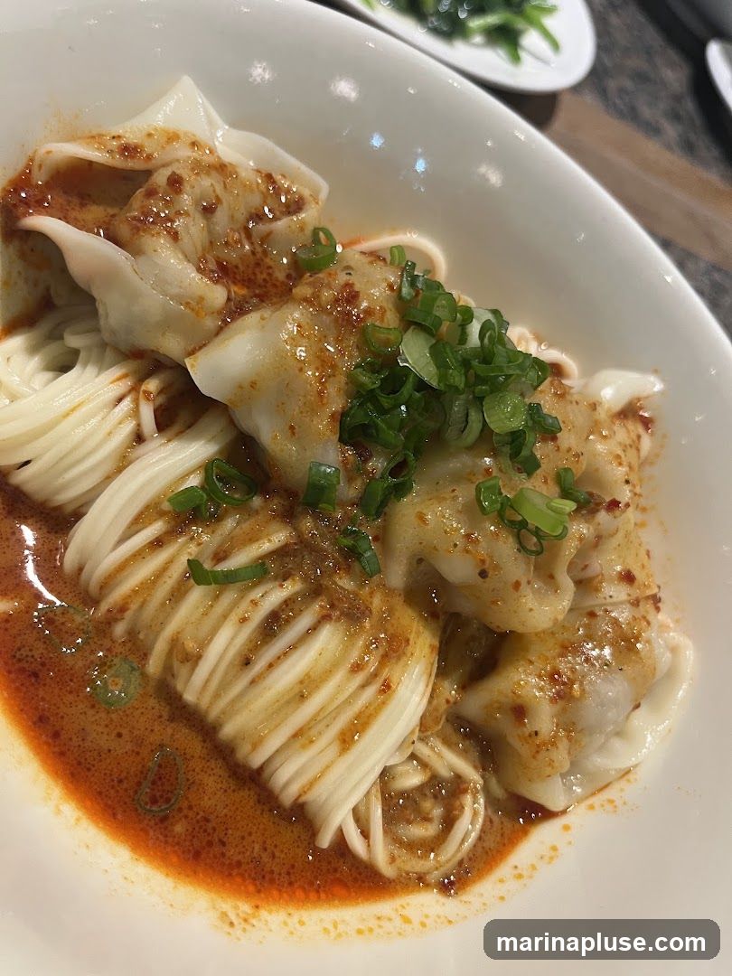 A close-up shot of a bowl of wontons and noodles in a spicy red sauce, garnished with chopped green onions.