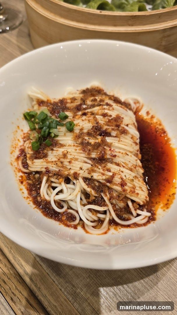A close-up shot of a bowl of white noodles, coated in a spicy chili oil sauce and garnished with chopped green onions. In the background, a bamboo steamer basket filled with green dumplings is visible. The dish is served on a wooden table.