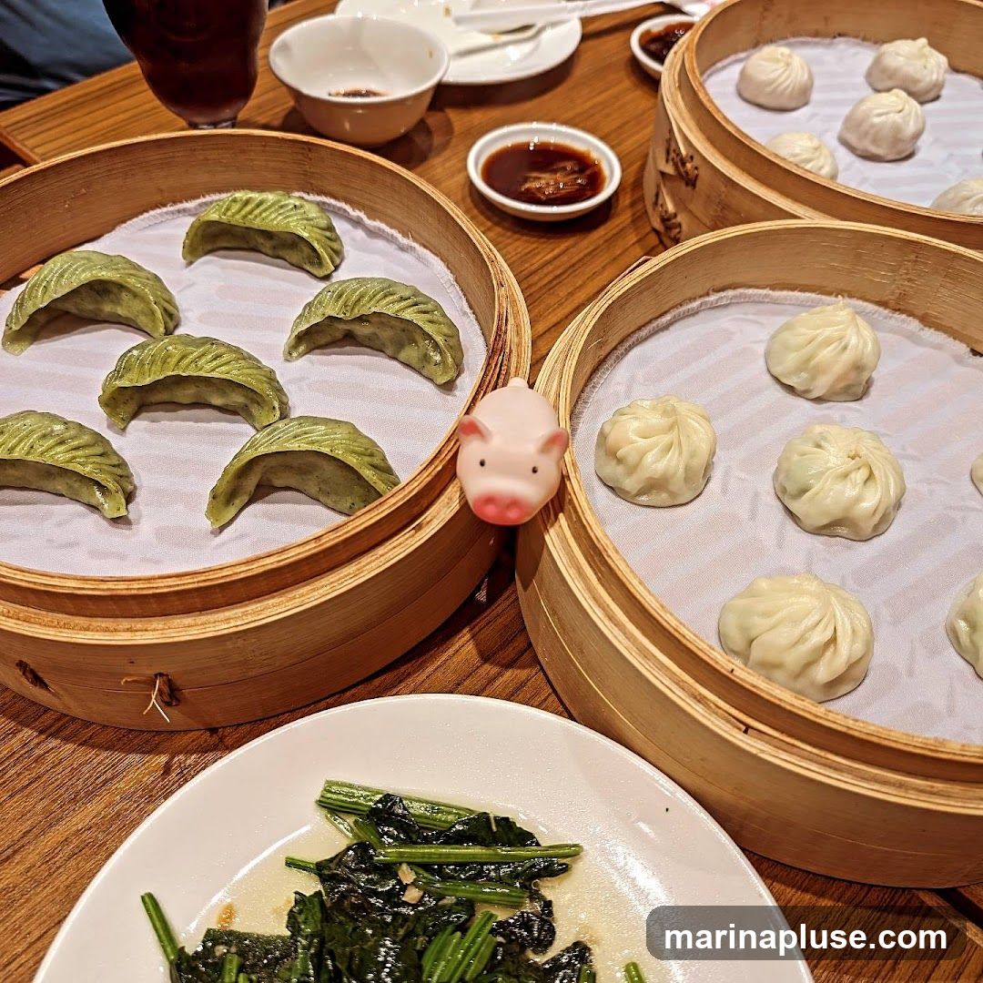 A close-up shot of a table laden with various dim sum dishes served in bamboo steamers. In the foreground, a plate of stir-fried greens is visible. The steamers contain green dumplings and soup dumplings, with a small pink pig figurine placed on one of the steamers. A dipping sauce and a drink are also present on the table.
