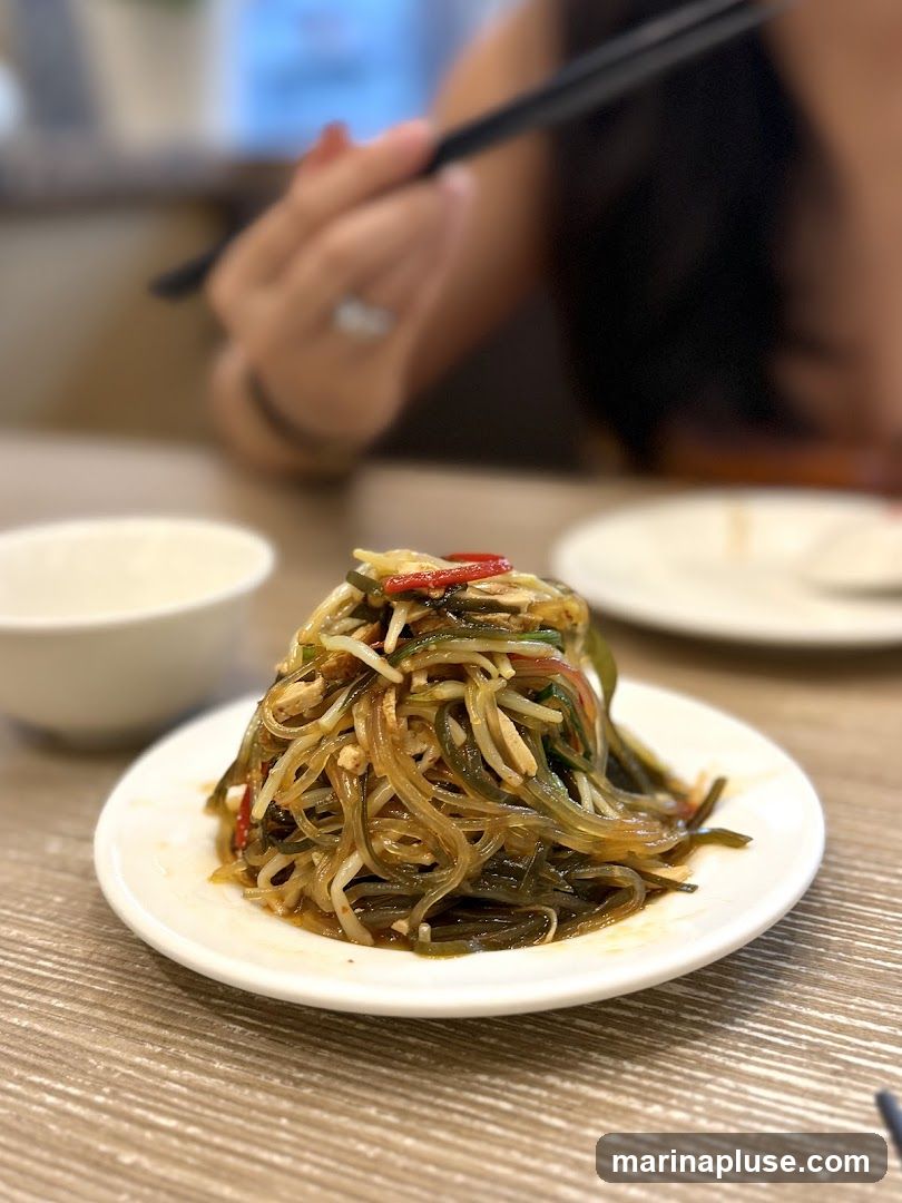 A close-up shot of a plate of noodle salad with a person in the background holding chopsticks. The salad appears to be made with glass noodles, seaweed, and possibly some shredded vegetables and meat, all tossed in a savory dressing. The dish is served on a white plate on a wooden table.
