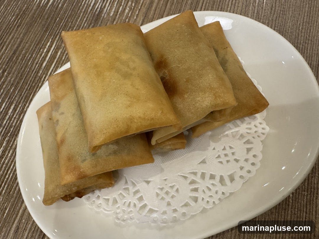 A plate of golden-brown spring rolls sits on a white plate with a decorative paper doily. The spring rolls are rectangular and appear to be fried to a crisp.