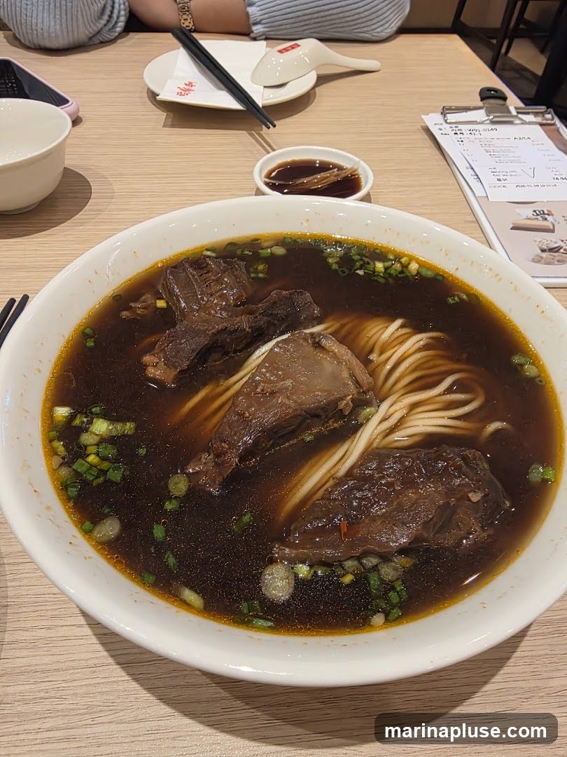 A close-up shot of a bowl of beef noodle soup, garnished with chopped green onions. The soup has a rich, dark broth and contains several large pieces of tender beef and noodles. A small dish of dipping sauce and chopsticks are visible on the side, along with a menu on a clipboard.