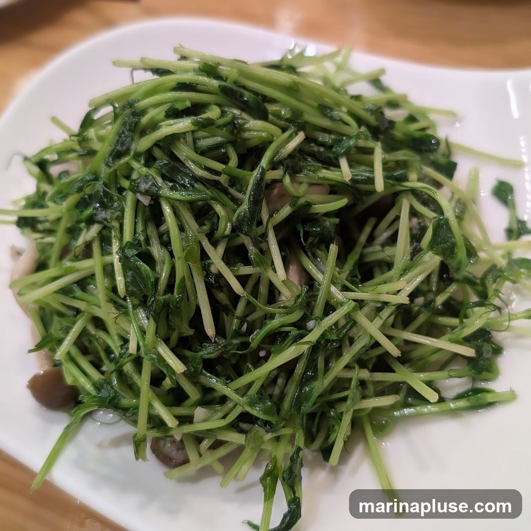 A close-up shot of a plate of stir-fried pea shoots, glistening with oil and mixed with what appear to be sliced mushrooms. The dish is served on a white, oval-shaped plate.