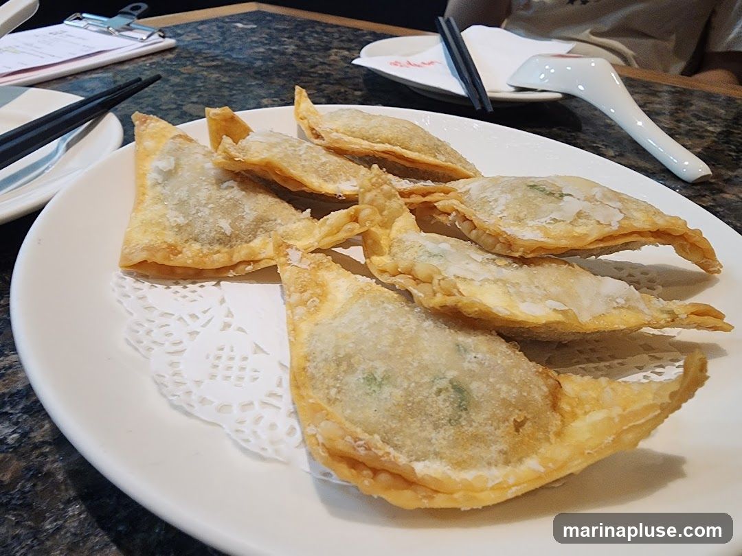 A plate of golden-brown fried wontons sits on a white doily on a white plate. The wontons are triangular in shape and appear to be filled with a green mixture. Chopsticks and a spoon are visible in the background, along with a menu or order pad.