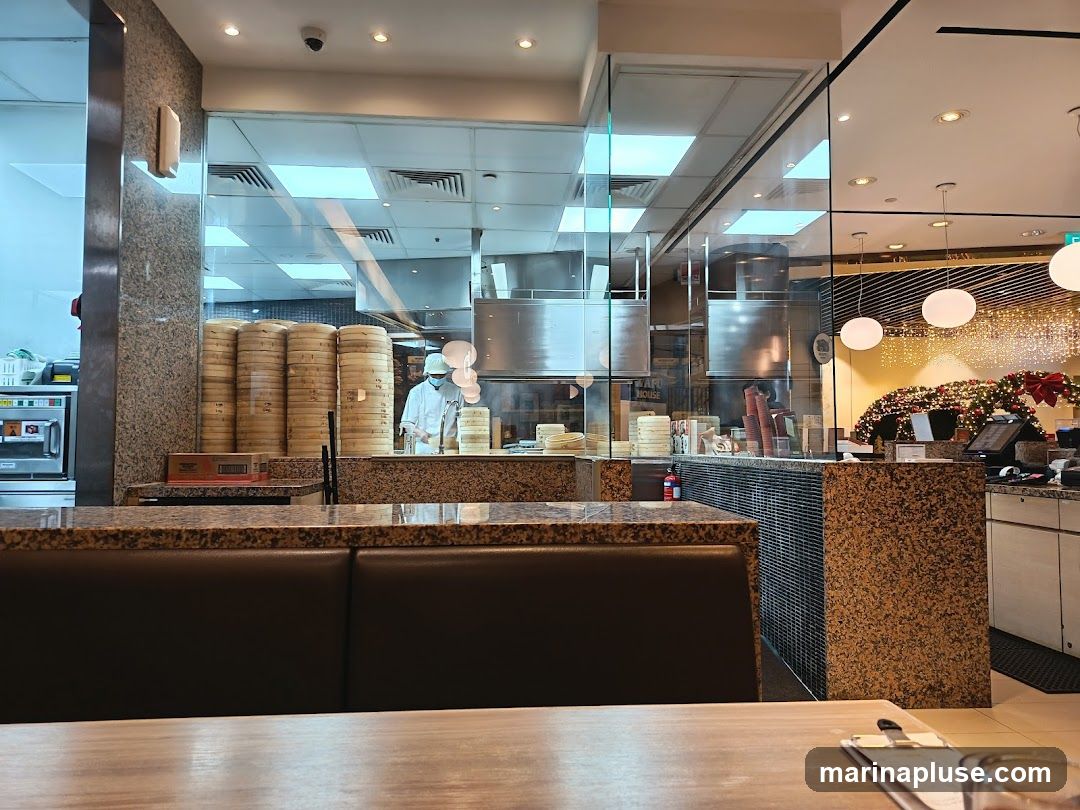 A view into a restaurant kitchen through a glass partition. Stacks of bamboo steamers are visible, along with a chef in a white uniform and mask preparing food. In the background, there are stainless steel kitchen appliances and ventilation hoods. The dining area features a counter with granite-like detailing, booth seating, and decorative holiday lights.