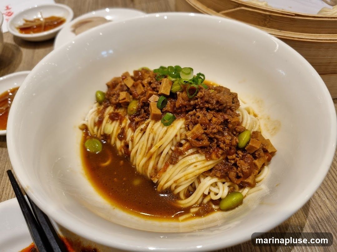 A close-up shot of a bowl of noodles topped with minced meat, edamame, and chopped green onions. The noodles are served in a savory sauce. In the background, there are small dishes with dipping sauce and a bamboo steamer.