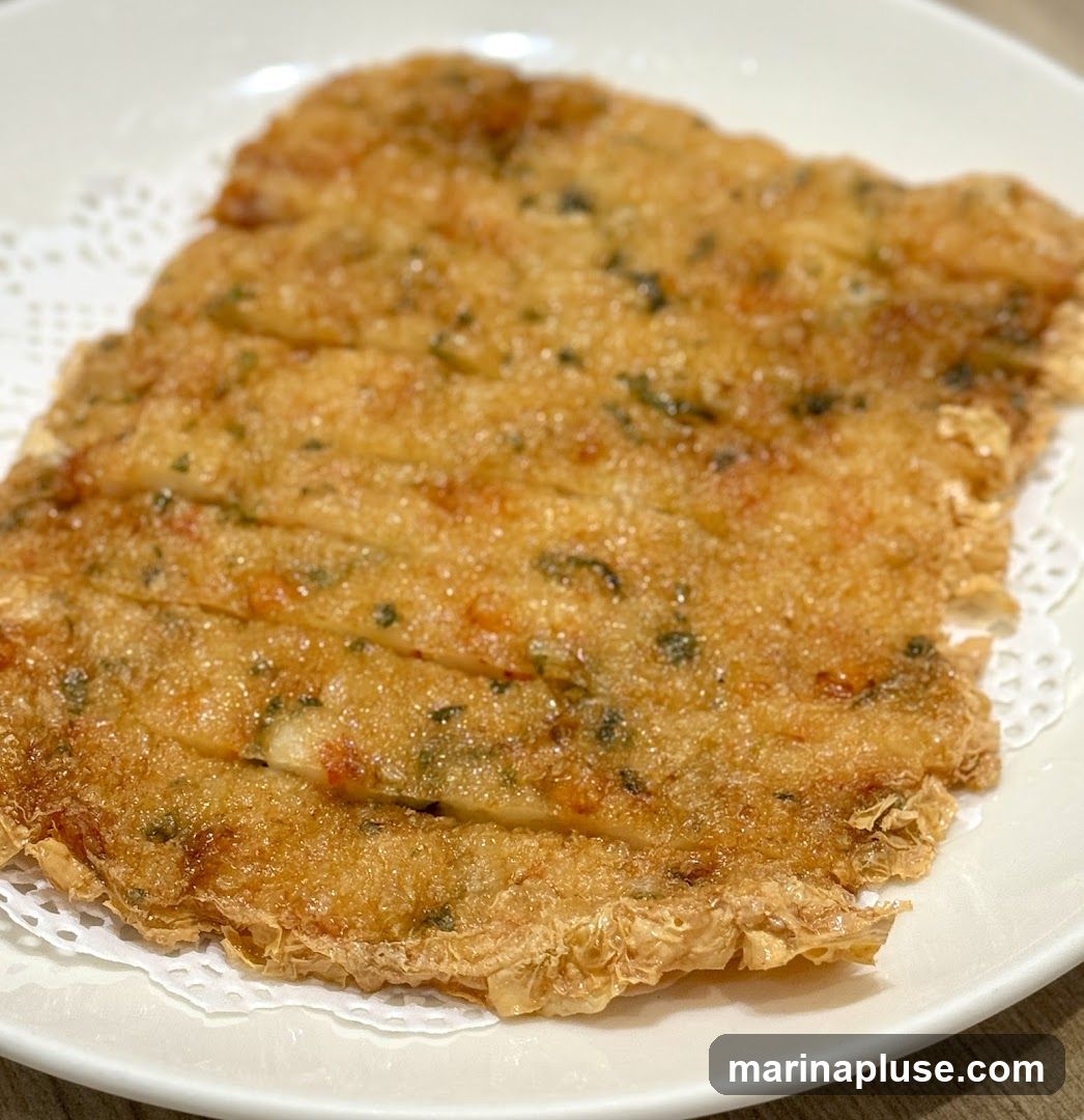 A close-up shot of a golden-brown fried bean curd skin roll, served on a white plate with a doily. The roll is cut into several sections and appears to be filled with chopped herbs and possibly small pieces of vegetables or seafood. The texture is crispy and layered.