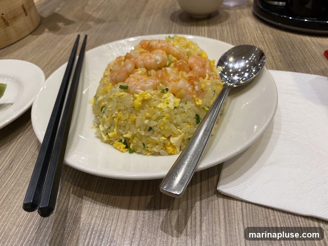 A plate of shrimp fried rice is served with chopsticks and a spoon on a wooden table. The fried rice is topped with several cooked shrimp and appears to contain scrambled eggs and green onions. A small white dish with a green condiment is visible to the left, and a white napkin is to the right. In the background, a bamboo steamer basket and a dark bowl are partially visible.