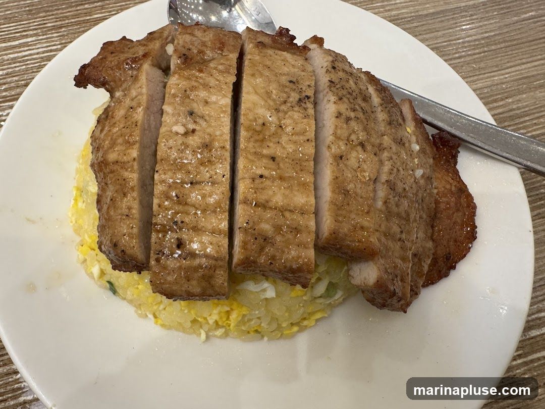 A close-up shot of a white plate filled with fried rice topped with sliced grilled pork chops. A spoon and a fork are visible on the side.